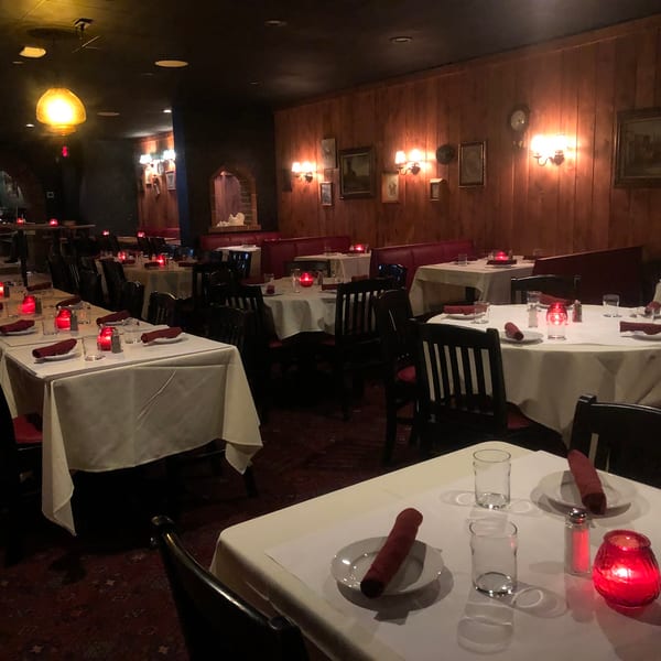 A dimly lit restaurant dining room with white-clothed tables, red accents, and wood-paneled walls.