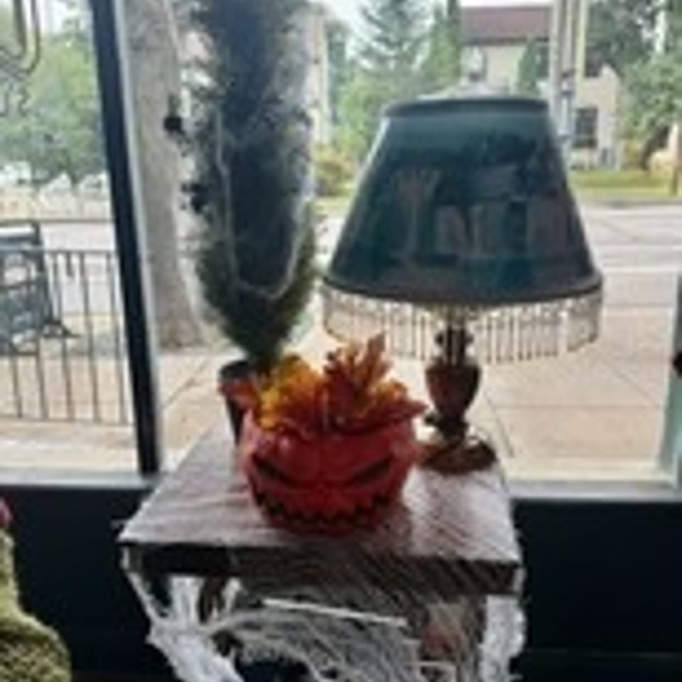 Ornate table lamp and red floral centerpiece sit on a lace-covered table beside a window.