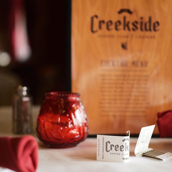 A Creekside Supper Club menu, matchbook, and red candle holder on a white tablecloth.
